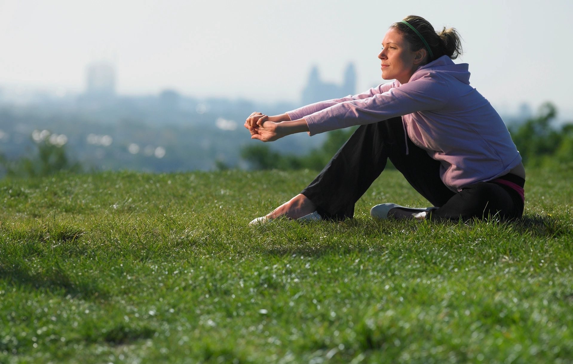 Woman sits in the grass, arms stretched out with her elbows on her knees.